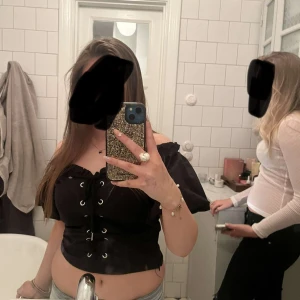Svart offshoulder korsettopp med snörning - Säljer en svart offshoulder korsettopp med snörning framtill och silverfärgade öljetter. Toppen är croppad och har korta ärmar som sitter snyggt över axlarna. Perfekt för dig som vill sticka ut med en edgy och trendig look. Lånade bilder!❤️