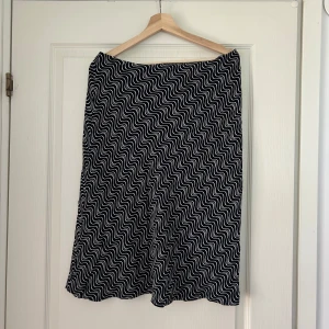 Svartvitt vågmönstrad kjol - Vintage lågmidjad midikjol från Ecoline med vitt vågigt mönster. Kjolen är knälång och har en rak, lätt figurnära passform. Mjuk och stretchig känsla, perfekt för dig som gillar grafiska prints och vill sticka ut lite extra.