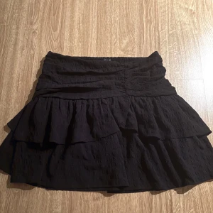 Svart volangkjol med struktur - Supersnygg svart kortkjol med volanger och strukturerat tyg. Kjolen har en hög midja och flera lager som ger en cool och trendig look. Perfekt att styla med en croppad topp eller oversized hoodie.