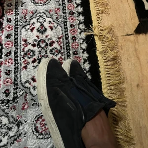 Svarta slip-on sneakers från Garment Project - Svarta slip-on sneakers från Garment Project med vit platt sula och elastiska band över vristen. Ovandelen är i mocka och skon har en stilren, minimalistisk look som passar till det mesta. Perfekta för dig som gillar enkel och clean stil.