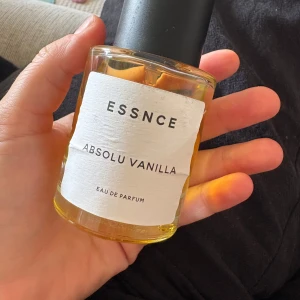 ESSNCE Absolu Vanilla parfym - En stilren parfymflaska från ESSNCE med doften Absolu Vanilla. Flaskan är rund och genomskinlig med svart lock och vit etikett. Innehållet är gyllengult och flaskan är i glas. Perfekt för dig som älskar varma, söta vaniljtoner.
