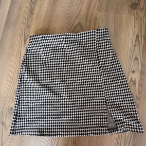 Svartvit rutig kortkjol med slits - Snygg svartvit kortkjol med klassiskt hundtandsmönster och liten slits framtill. Kjolen har en rak passform och är perfekt för dig som gillar en stilren men ändå trendig look. Passar lika bra till en stickad tröja som till en cool t-shirt.