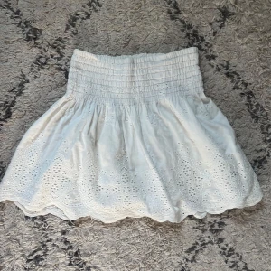 Vit kortkjol med broderi By Malina - Supersöt vit kortkjol från By Malina med bred smock i midjan och broderade detaljer längs fållen. Kjolen har volangig nederkant och är gjord i luftig bomull, perfekt för varma dagar. 