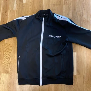 Palm Angels kofta - Svart track jacket från Palm Angels med vita ränder längs ärmarna och vit dragkedja. Jackan har ståkrage, två fickor med dragkedja och logga på bröstet. Materialet är en mix av bomull och polyester för en sportig vibe.