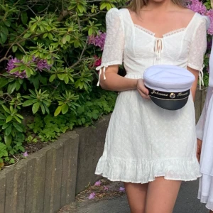 Vit kort klänning med broderi och volang - Supersöt vit kort klänning med broderade blommor, puffärmar och knytning fram. Klänningen har volangkant nertill och romantiska detaljer som spets vid urringningen. Perfekt för sommarens alla tillfällen!