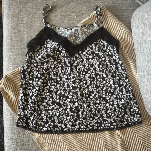 Svart blommig linnetopp från Amisu - Supersöt svart linnetopp från Amisu med vita blommor och tunna axelband. Toppen har spetsdetaljer vid urringningen och är perfekt för en chill och trendig look. Lätt och luftig, passar perfekt till jeans eller kjol.