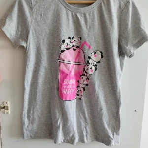 Grå t-shirt med panda-tryck - Söt grå t-shirt med korta ärmar och ett stort tryck av glada pandor och en rosa dryck med texten 'Sunny and Happy' på framsidan. Perfekt för dig som gillar gulliga motiv och vill ha en avslappnad stil.Kan hämtas upp i Malmö 