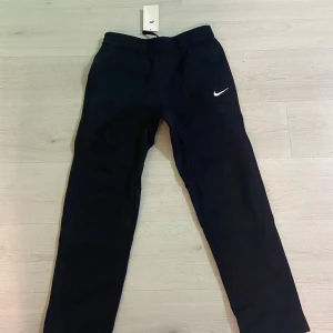 Helt nya Svarta mjukisbyxor från Nike - Helt nya Svarta mjukisbyxor från Nike med klassisk vit logga på låret. Byxorna har resår i midjan och är tillverkade i mjukt bomullsmaterial. Perfekta för chill eller träning, med en slim passform som sitter snyggt.