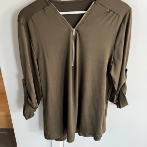 Olivgrön långärmad tröja med dragkedja - Olivgrön långärmad tröja i 100% bomull med snygg v-ringning och silverfärgad dragkedja framtill. Ärmarna kan rullas upp och fästas med knapp för en mer avslappnad look. Perfekt för dig som gillar stilrena och bekväma plagg.