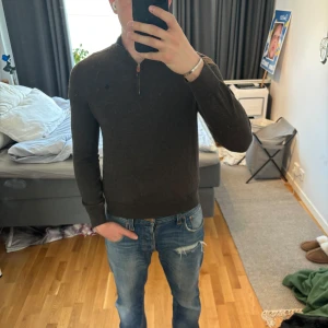 Mörkgrå merinoullströja Morris M - Snygg mörkbrun stickad tröja från Morris i 100% merinoull. Tröjan har en stilren half zip-dragkedja vid halsen och diskret broderad logga på bröstet. Perfekt för dig som gillar klassisk och clean stil med en modern touch. Storlek M