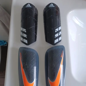 Adidas och Nike benskydd fotboll - Två par benskydd för fotboll, ett från Adidas i svart med vita detaljer och geometriskt mönster, samt ett från Nike i mörkgrått med orange swoosh. Båda är formade för att sitta bekvämt på smalbenet och har en skyddande yta i plast.