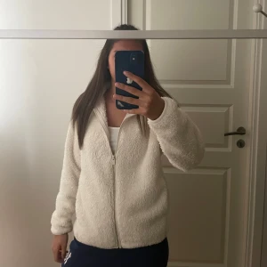Vit teddyjacka från Uniqlo, S - Supermjuk och fluffig vit teddyjacka från Uniqlo i storlek S. Jackan har dragkedja framtill, hög krage och är helfärgad utan mönster. Perfekt för kyliga dagar när du vill vara både varm och stilren. Den kan även användas som en kofta under milda sommarkvällar. Storleken är i S men passar även dig som bär på Xs.