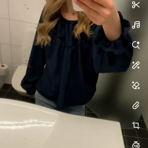 Mörkblå blus med volangärm - Säljer en mörkblå blus med snygga volangärmar och lös passform. Blusen har rundad halsringning och är tillverkad i ett lätt, strukturerat tyg som känns luftigt. Perfekt för dig som gillar en stilren men ändå trendig look.