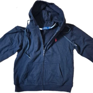 Svart hoodie från Polo Ralph Lauren - Snygg svart hoodie från Polo Ralph Lauren med dragkedja och huva. Klassisk röd broderad logga på bröstet och stora fickor framtill. Perfekt för en chill och stilren look. Tillverkad i mjuk bomull för extra komfort.