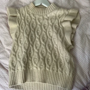 Beige stickad väst med volang från Name It - Supersöt beige stickad väst från Name It i storlek 116. Västen har ett snyggt kabelstickat mönster och breda ribbade kanter. Ärmarna har volangdetaljer som ger en extra touch. Perfekt att styla över en skjorta eller t-shirt för en trendig look.