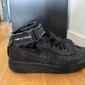 Nike Air Force 1 Mid Comme Des Garcons Black - Svarta Nike Air Force 1 mid x Comme des Garçons. Skorna är i skinn med snörning och kardborreband upptill, där Comme des Garçons-loggan syns.  Säljs på stockx för ca 5000 kr (se sista bilden) , jag kan gå ner från 2400 (mitt pris) vid snabb affär! 