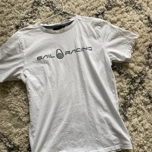 Vit t-shirt från Sail Racing - Vit t-shirt från Sail Racing med svart logotyptryck över bröstet. Klassisk rund halsringning och korta ärmar. Tillverkad i mjuk bomull, perfekt för en clean och sportig look.