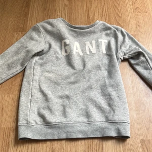 Grå sweatshirt från GANT - Snygg grå sweatshirt från GANT med stora vita bokstäver framtill. Klassisk rund hals och långa ärmar. Mjuk och skön bomullsblandning som passar perfekt till jeans eller joggers. Enkel och stilren design för en chill vardagslook.