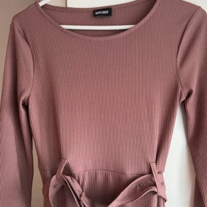 Ribbad lila långklänning med knyt - Säljer en ribbad lila långklänning från Missguided med långa ärmar och snygg knytning i midjan. Klänningen har en enkel, rak passform och rund halsringning. Perfekt för dig som gillar stilrena och bekväma plagg.