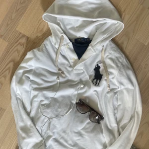 Vit big pony hoodie från Ralph Lauren.  - Vit hoodie från Polo Ralph Lauren med stor svart logga på bröstet. Tröjan har huva med snörning, knappar vid halsen och är tillverkad i bomull. Långärmad och klassisk passform, perfekt för en clean och stilren look. Storlek S i bra skick utan defekter. 