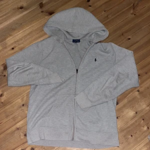 Grå hoodie från Ralph Lauren - Snygg ljusgrå hoodie från Ralph Lauren med dragkedja och klassisk huva. Tröjan har lång ärm och den ikoniska lilla loggan broderad på bröstet. Perfekt för chill dagar eller när du vill ha en enkel och stilren look.