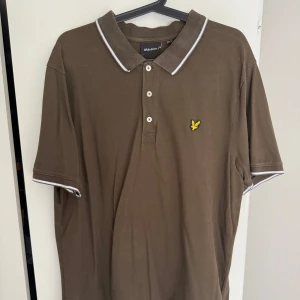 Oliven  pikétröja från Lyle & Scott xxl - Snygg oliven pikétröja från Lyle & Scott med klassisk krage, tre knappar och vit kant på krage och ärmslut. Ikonisk gul logga broderad på bröstet. Perfekt för en stilren och avslappnad look.