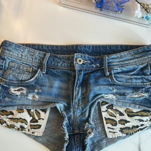 Jeansshorts med leoparddetaljer H&M - Snygga blå jeansshorts från H&M med slitna detaljer och råa kanter. Insidan av fickorna har coolt leopardmönster i vitt, svart och guld. Klassisk femficksmodell med låg midja och &Denim-lapp bak. Perfekta för en edgy sommarlook!