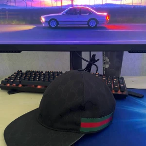 Svart keps från Gucci med logomönster - Svart keps från Gucci med diskret GG-logomönster över hela kepsen och klassiska gröna och röda ränder på sidan. Snygg och stilren modell med böjd skärm, perfekt för att lyfta din outfit.Ny men andvänd.