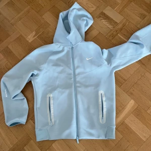 Nike NOCTA ljusblå vindjacka med huva - Nike NOCTA vindjacka i ljusblått med hel dragkedja, huva och två fickor med dragkedja framtill. Jackan har snygga detaljer som NOCTA-tryck på ärmen och diskret Nike-logga på bröstet. Perfekt för dig som gillar streetwear och sportig stil. Tveka inte på att höra av er vid frågor ganska stor i storleken också. Den är helt oanvänd.