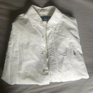 Vit skjorta från Ralph Lauren Slim Fit - Klassisk vit skjorta från Ralph Lauren i slim fit-modell. Skjortan har knappar framtill, button down-krage och den ikoniska broderade polospelaren på bröstet. Tillverkad i mjuk bomull, perfekt för en clean och stilren look.