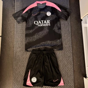 PSG x Nike svart fotbollströja M - Säljer en svart PSG fotbollströja från Nike i storlek M. Tröjan har korta ärmar, rosa detaljer på axlarna och ett grått mönster över hela framsidan. Materialet är lätt och andas, perfekt för träning eller match. Qatar Airways-logga och PSG-emblem på bröstet.