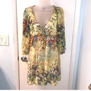 Blommig kortklänning med snörning - Superfin kortklänning med blommigt mönster i ljusa färger som gul, beige, blå och röd. Djup v-ringning med snörning framtill och långa, transparenta ärmar med resår vid handlederna. Flowy passform och lätt material, perfekt för vår och sommar.