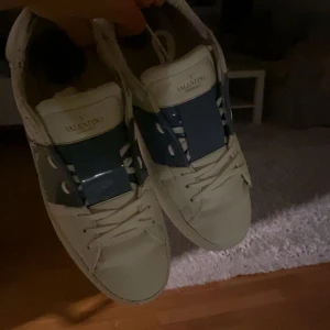 Valentino Garavani Open sneakers vit/blå - Säljer ett par Valentino Garavani Open sneakers i vitt skinn med bred blå rem över vristen och vita snören. Klassisk rund tå och platt sula, riktigt snygga och stilrena med exklusiv känsla. Kommer med originalkartong och extra skosnören. Självklart äkta och äktehetsbevis medföljer vid köp.