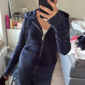 Mörkblå velourjacka Juicy Couture XXS - Mörkblå velourjacka från Juicy Couture med huva och dragkedja framtill. Tröjan har långa ärmar, fickor på sidorna och klassisk broderad logga på bröstet. Perfekt för en chill och trendig look.