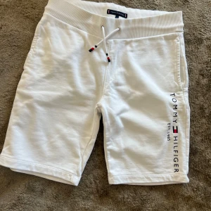 Vita shorts från Tommy Hilfiger - Snygga vita shorts från Tommy Hilfiger med bred ribbad midja och snörning framtill. Mjukt bomullsmaterial och logga tryckt längs ena benet. Bakficka med broderad flagga. Perfekta för sommardagar och chill stunder. Strl 152. Inga fläckar - fint skick! Lite använda 