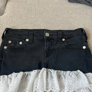 Jeanskjol med spets från True Religion - Supersnygg svart jeanskjol från True Religion med vit volangspets. Är inte använd men lappen är inte kvar.