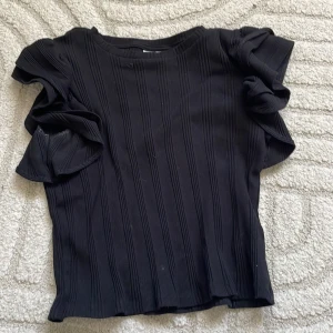 Svart ribbad topp från Gina Tricot - Svart ribbad topp från Gina Tricot med korta volangärmar. Toppen har en rund halsringning och är tillverkad i ett stretchigt material som sitter snyggt på kroppen. Perfekt för dig som gillar en stilren look med en twist.