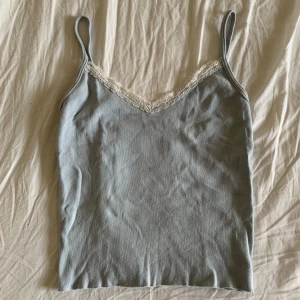 Ljusblå linnetopp med spetskant - Supersöt ljusblå linnetopp med tunna axelband och v-ringning dekorerad med vit spets. Perfekt att styla med jeans eller kjol för en chill look. Mjuk och skön känsla, enkel att matcha till olika outfits.