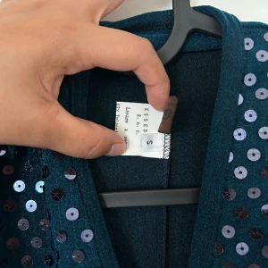 Mörkblå paljettklänning från Essedue - Supersnygg mörkblå klänning med lång ärm och v-ringning. Framsidan är täckt av glittriga paljetter som ger en festlig vibe. Klänningen har en omlott-look upptill och är figurnära upptill med markerad midja. Perfekt för dig som vill sticka ut.