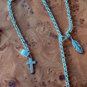 Silverfärgat halsband med kors och fjäder - Kedjehalsband i silverfärg med två hängen: ett kors och en detaljerad fjäder. Kedjan är grov och ger en cool vibe. Ett extra hänge i guld med fjädermotiv ingår också. Perfekt för dig som gillar att mixa symboler och stilar.