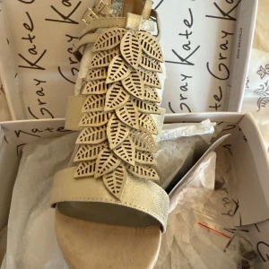 Guldiga sandaler med klack från Kate Gray - Snygga sandaler i guldigt skinn med hög klack och öppen tå. Dekorerade med detaljerade blad i flera lager framtill och justerbart spänne vid ankeln. Perfekta för dig som vill sticka ut med en unik och elegant look.