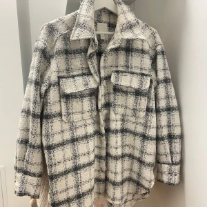 Rutig overshirt från H&M i ullmix - Snygg rutig overshirt från H&M i svart och vit ullmix. Jackan har stora bröstfickor med lock, knappar framtill och klassisk krage. Perfekt lager-på-lager-plagg med avslappnad passform och trendig look.