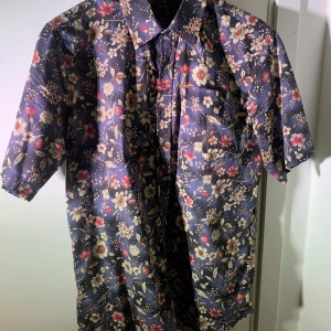 Blommig kortärmad skjorta från Berga Storlek M.  - Snygg kortärmad skjorta från Berga i marinblått med blommigt mönster i beige, rött och vitt. Skjortan har klassisk krage, bröstficka och knäppning framtill. Perfekt för dig som vill sticka ut med en färgglad och trendig look.