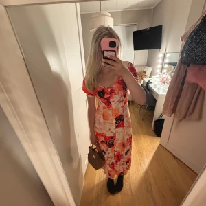 Blommig långklänning - Säljer en färgstark långklänning med stora blommor i rött, orange, svart och vitt. Klänningen har korta puffärmar, söt rynkad urringning och knappar framtill. Priset går att diskutera kontakta mig vid frågor !❣️