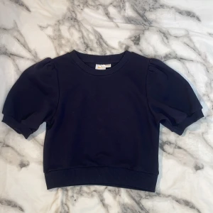 Mörkblå puffärm tröja från Gina Tricot - Kortärmad mörkblå tröja från Gina Tricot med trendiga puffärmar och ribbade muddar vid ärmslut och nederkant. Tillverkad i mjuk bomull, perfekt för en stilren och enkel look. Snygg att matcha med både jeans och kjol.