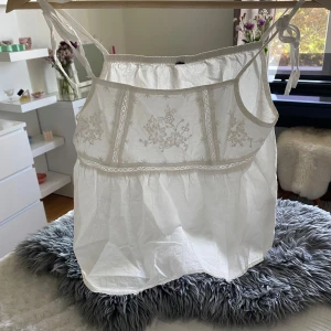 Vit broderad linnetopp med knytband - Supersöt vit linnetopp med tunna axelband som knyts på axlarna. Toppen har broderade blommor framtill och volang nedtill för en romantisk vibe. Perfekt för varma dagar och lätt att matcha med jeans eller kjol.