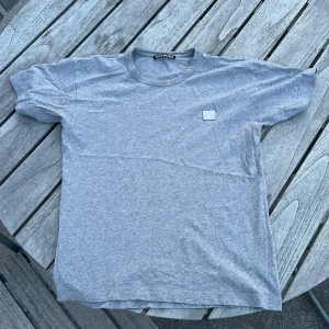 Grå t-shirt från Acne Studios - Snygg grå t-shirt från Acne Studios med klassisk passform och rund hals. T-shirten har korta ärmar och den ikoniska Acne Studios-loggan på bröstet. Perfekt basic-plagg i mjuk bomull som funkar till allt och speciellt till sommaren. Tröjan är mycket i ett mycket fint skick. 