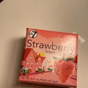 W7 Strawberry Street Rouge - Rouge från W7 i färgen Strawberry Street. Kommer i en söt rosa förpackning med jordgubbsdesign och illustration av hus. Perfekt för att ge kinderna en fräsch, bärig ton. Innehåller 6g puderrouge.