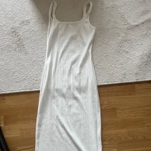 Vit ribbad långklänning - Snygg vit långklänning med ribbad struktur och smala axelband. Klänningen har en enkel och stilren design som sitter figurnära och är perfekt för varma dagar. Passar dig som gillar minimalistisk stil och vill ha något bekvämt men ändå trendigt.