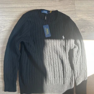 Svart kabelstickad tröja Polo Ralph Lauren - Svart kabelstickad tröja från Polo Ralph Lauren med klassisk rund halsringning och broderad vit logga på bröstet. Tröjan har lång ärm och ribbade muddar. Perfekt för dig som gillar stilren och tidlös design.Den är lite liten i storlek.